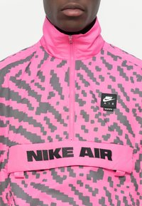 Nike Air jacket featuring a pink and gray pixelated pattern, with a high collar, half-zip closure, and a black branded waistband.