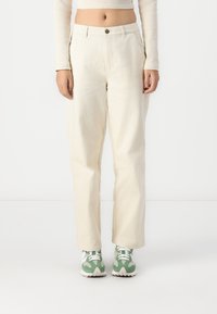 Hellbeige weite Hosen aus Baumwolle mit einem vorderen Knopfverschluss und Seitentaschen, kombiniert mit grünen und weißen Sneakers.