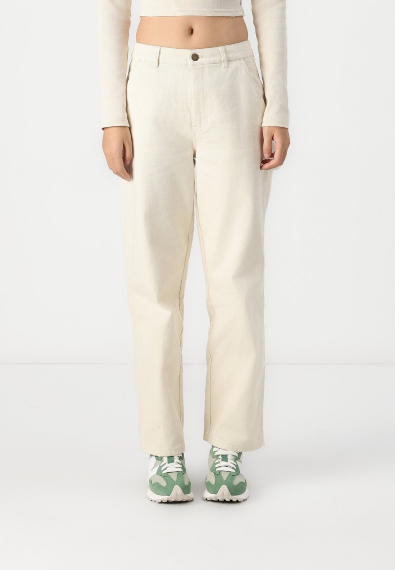 Hellbeige weite Hosen aus Baumwolle mit einem vorderen Knopfverschluss und Seitentaschen, kombiniert mit grünen und weißen Sneakers.