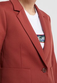 Blazer couleur rouille avec une coupe structurée, revers crantés, fermeture à un bouton et une poche avant. Porté sur un t-shirt blanc à motif.