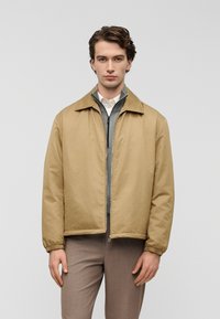 Ελαφρώς καφέ, υφασμένη ζακέτα με γιακά και ελαστικά μανσέτες, φορεμένη πάνω από γκρι μπλούζα με φερμουάρ. Το μοντέλο φοράει ελαφρώς καφέ παντελόνια.