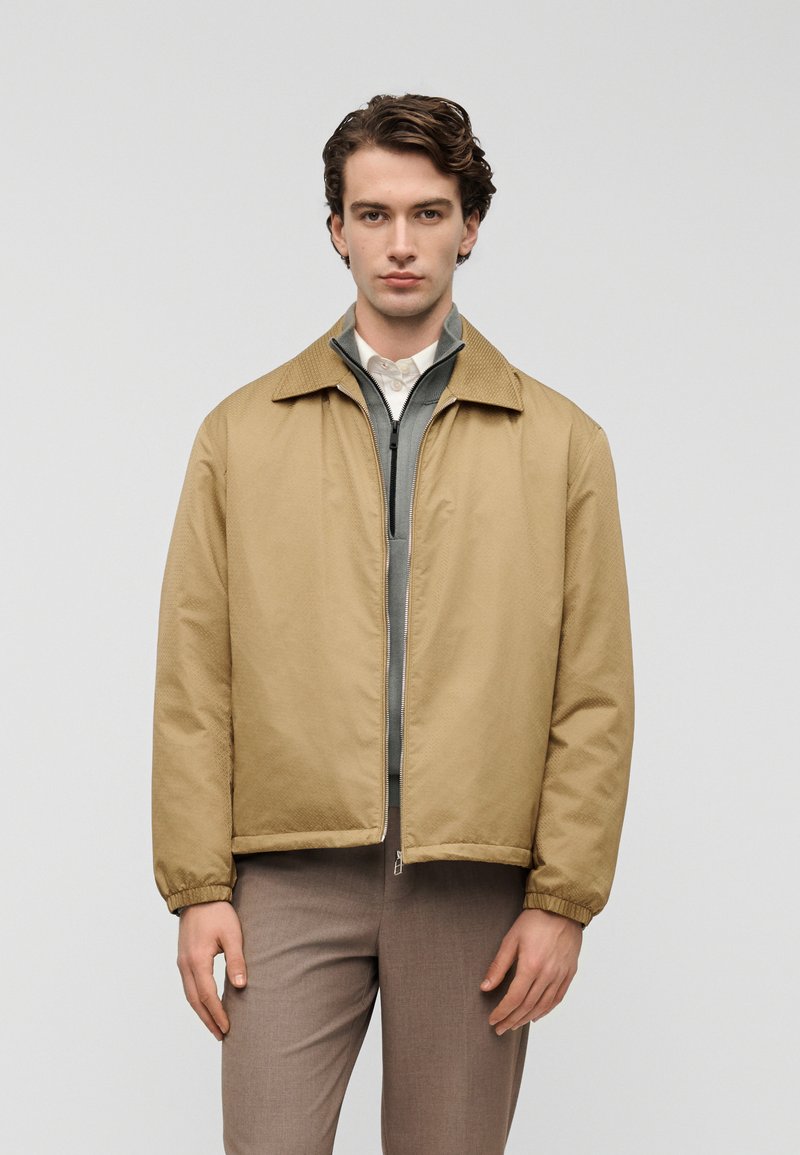 Ελαφρώς καφέ, υφασμένη ζακέτα με γιακά και ελαστικά μανσέτες, φορεμένη πάνω από γκρι μπλούζα με φερμουάρ. Το μοντέλο φοράει ελαφρώς καφέ παντελόνια.