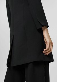 Schwarze Blazer mit langen Ärmeln und seitlichem Schlitz, der über einer passenden schwarzen Hose getragen wird, von hinten gezeigt mit einer entspannten Hand an der Seite.