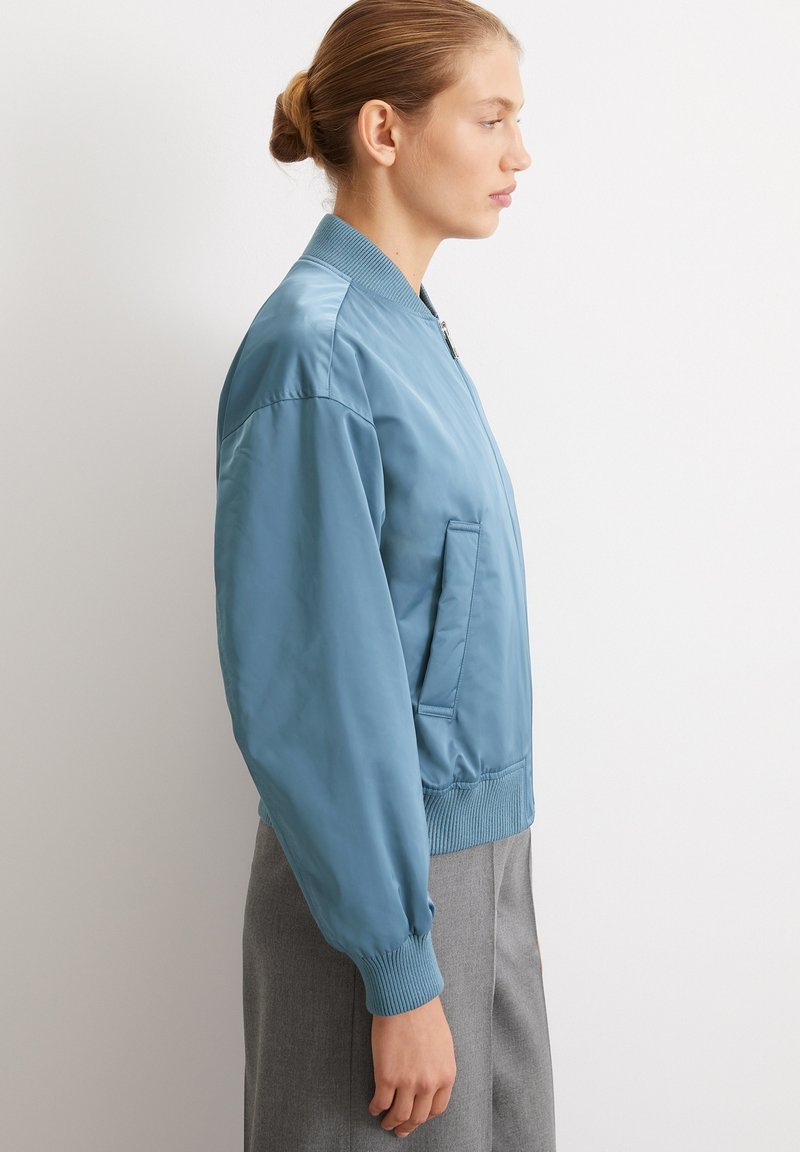 Jasnoniebieska kurtka bomberka wykonana z gładkiego, błyszczącego materiału, z okrągłym kołnierzem, ściągaczami na mankietach i boczną kieszenią. Dopasowany krój.