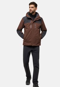 Giacca impermeabile marrone e grigia con cappuccio, cerniere e tasche. Abbinata a pantaloni neri e scarpe, con una vestibilità su misura.