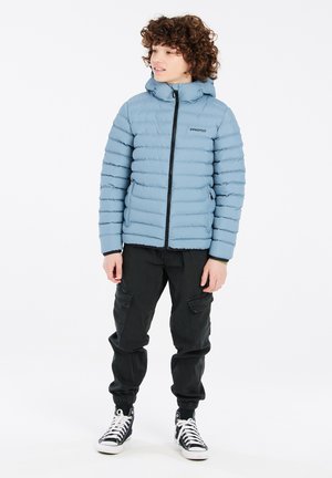 Lichtblauwe gewatteerde jas met horizontale quilting, zwarte rits en capuchon. Gecombineerd met zwarte cargobroek en hoge sneakers.