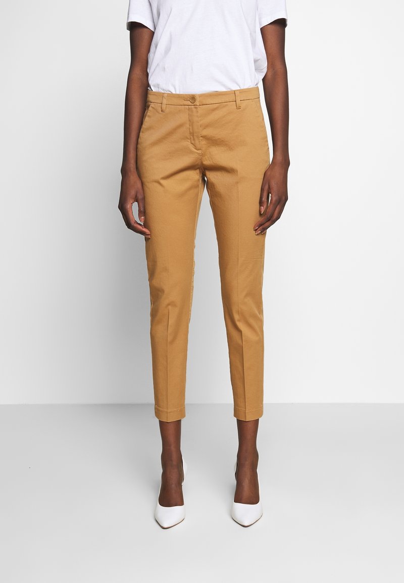 Pantalon en coton beige avec une coupe ajustée, longueur raccourcie et devant plat. Porté avec des talons blancs à bout pointu.