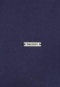 Pequeña etiqueta rectangular de metal plateado con texto en negro "SMILODOX" cosida sobre tela de ribete azul oscuro.