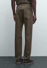 Person wearing straight-leg olive green pants and dark brown casual shoes, standing against a plain light gray background.