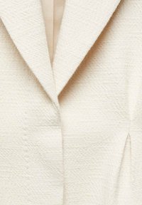 Blazer v kremni barvi, ki se ponaša z teksturirano tkanino, strukturiranim ovratnikom in zapiralom na en gumb. Prilagojen kroj z elegantnimi šivi.