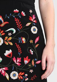 Černá tkanina vyšívaná barevnými červenými, bílými, modrými a žlutými květy, ukázaná vedle pravé ruky a předloktí osoby.