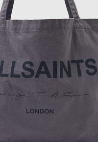 Pilka drobų rankinė su dideliu juodu „ALLSAINTS“ logotipu ir mažesniu tekstu, turi tvirtas rankenas ir tekstūruotą paviršių.