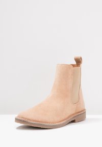 Bota Chelsea de ante beige con paneles laterales elásticos, suela de goma plana marrón, puntera redondeada y un tirador en el talón para facilitar su uso.