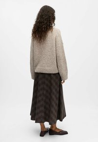Pull en laine brun clair avec une finition texturée, associé à une longue jupe écossaise brun foncé avec un motif en chevrons et des chaussures plates marron.