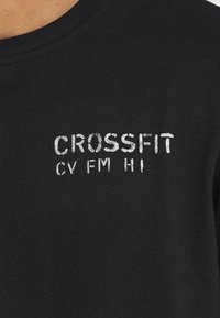 Černé bavlněné tričko s bílým potiskem textu "CROSSFIT" a "CV FM HI" na hrudi, kulatým výstřihem a krátkými rukávy.