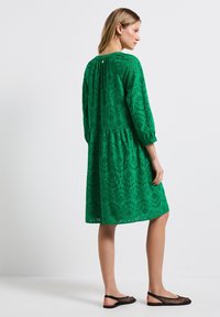 Abito verde in cotone con ritagli ricamati, orlo peplum e maniche a tre quarti. Chiusura a chiave sulla schiena. Indossato con ballerine in rete nera.