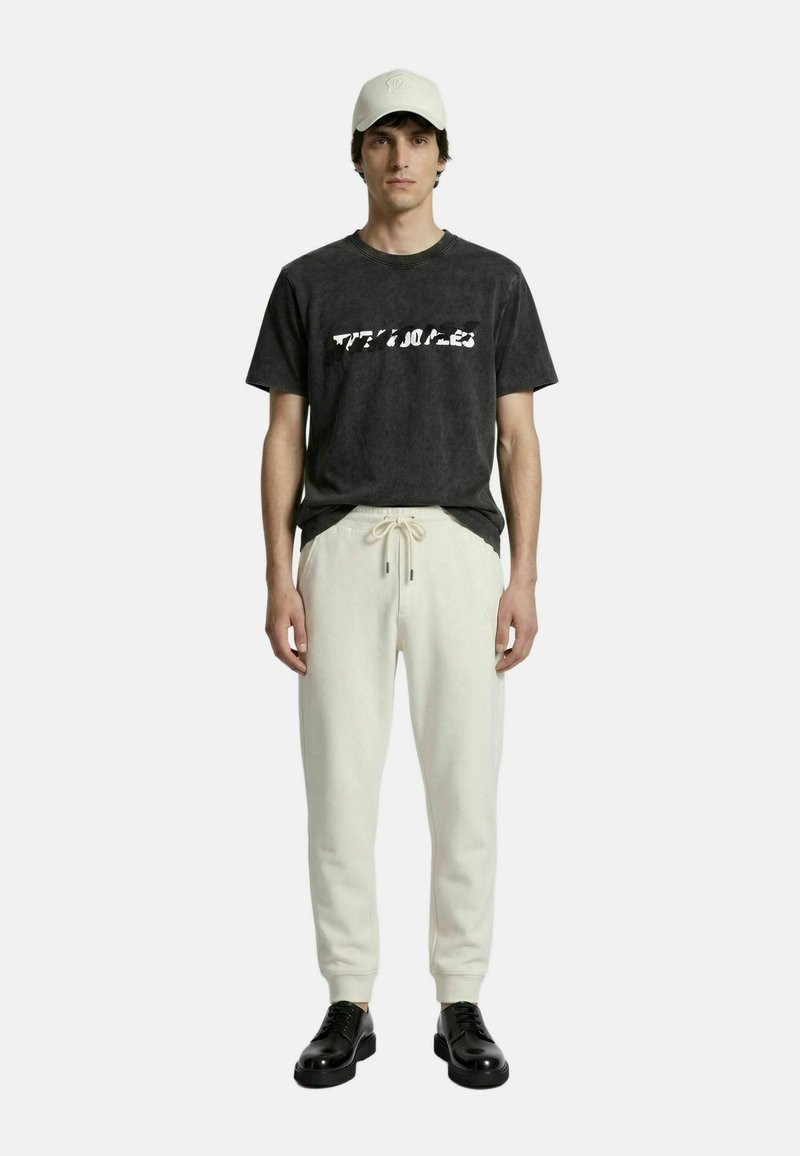 Hombre con camiseta negra con diseño gráfico, pantalones deportivos crema con cordón, zapatos negros y gorra de béisbol clara, de pie frente a un fondo liso.