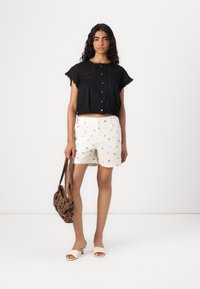 Blusa nera cropped con dettagli in pizzo, pantaloni corti bianchi con motivi floreali colorati, mule beige con tacco e una borsa marrone.