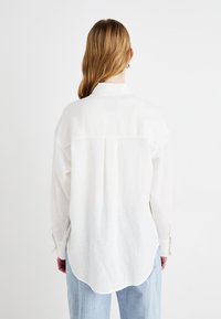 Chemise en lin blanc avec col, épaules tombantes et ourlet incurvé. Les manches longues présentent des poignets boutonnés. Texture douce et légère.