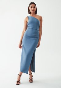 Blaues, einseitig geschnittenes Maxikleid aus glattem, dehnbarem Stoff mit einem seitlichen Schlitz und Raffung, kombiniert mit schwarzen Riemchen-High-Heels.