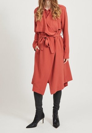 Femme portant un long manteau ceinturé couleur rouille sur un pantalon noir et des bottines noires à talons hauts pointues, debout devant un fond uni.