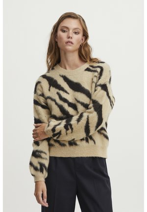 Un pull beige avec des rayures de tigre noires, fabriqué dans un matériau doux. Il présente un col rond et des poignets côtelés, porté par-dessus un pantalon sombre.