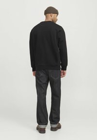 Felpa nera con polsini a coste, abbinata a jeans scuri con orlo arrotolato e cuciture a contrasto. Un berretto marrone completa il look.