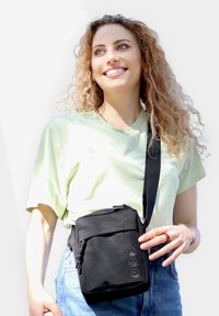 Borsa a tracolla nera in tessuto con tracolla regolabile, dotata di una tasca con zip e tre loghi sulla parte frontale, indossata su una camicia verde chiaro.