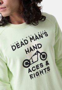 Sudadera verde claro con texto impreso en negro y un gráfico de bicicleta. Cuello redondo y puños de canalé. Textura de tela suave.