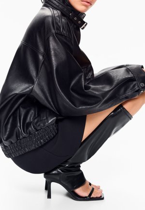 Žena v podřepu, oblečená v nadměrné černé kožené bundě, černé mini sukni a černých botách na podpatku s otevřenou špičkou.