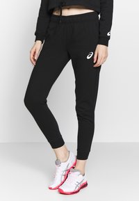 Zwarte joggingsbroek met elastische tailleband, geribbelde boorden en Asics-logo detail. Gepaard met witte en roze sport schoenen.