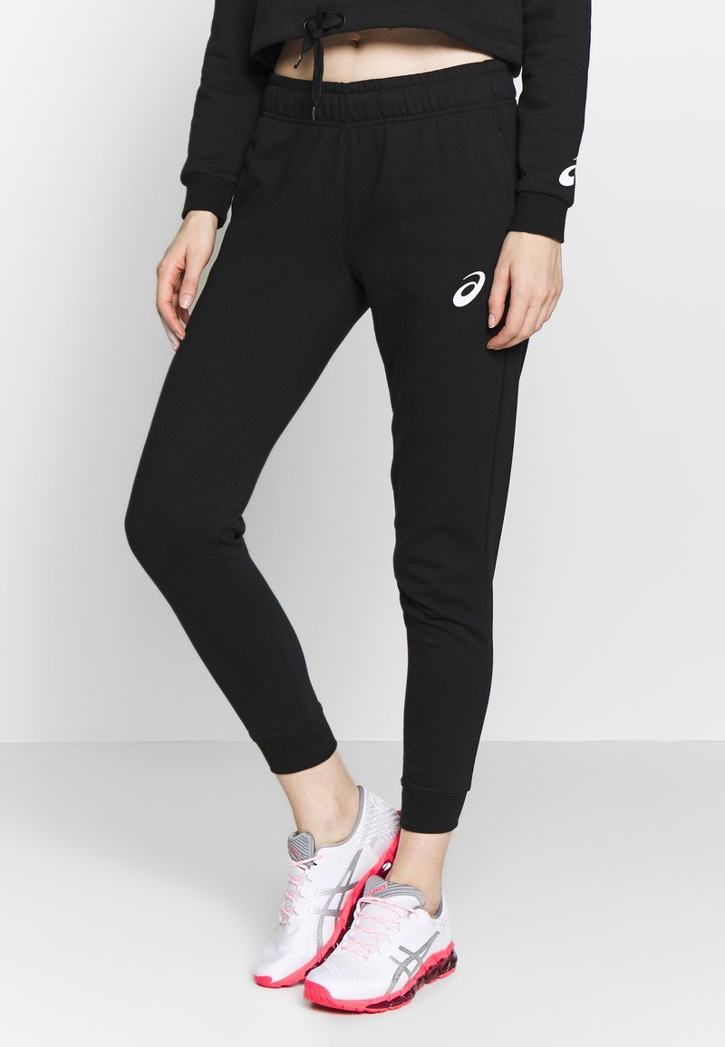 Zwarte joggingsbroek met elastische tailleband, geribbelde boorden en Asics-logo detail. Gepaard met witte en roze sport schoenen.