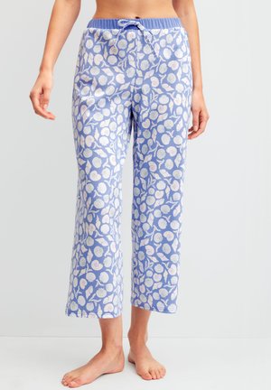 Vrouw die lichtblauwe pyjamabroek draagt met patroon van witte schelpen en koraal, staat blootvoets tegen een effen achtergrond.