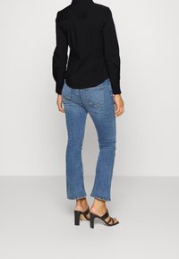 Camisa negra de botones con mangas largas, combinada con jeans de color azul claro y sandalias de tacón alto negras de dedo abierto. Costuras sutiles en los jeans.