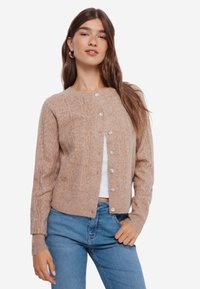 Beige gebreide cardigan met een ronde halslijn en knoopsluiting, voorzien van een gemêleerd patroon en geribde manchetten. Gekocht over een witte top.