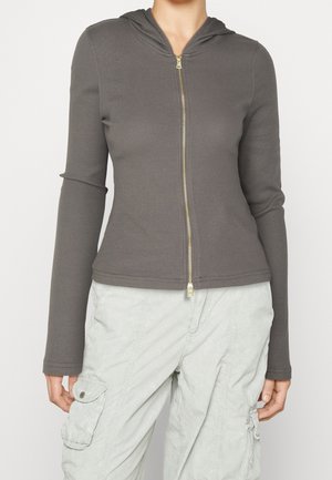 Femme portant une veste à capuche gris ajustée à fermeture éclair avec une texture côtelée et un pantalon cargo gris clair avec poches latérales.