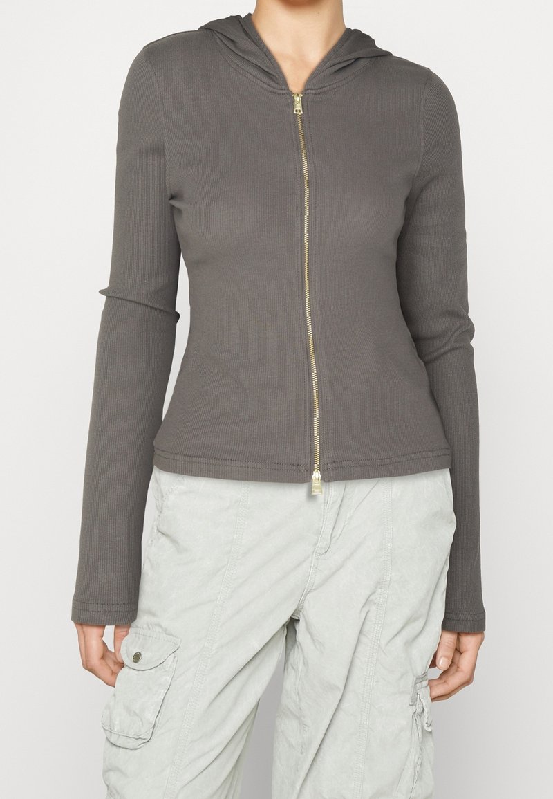 Femme portant une veste à capuche gris ajustée à fermeture éclair avec une texture côtelée et un pantalon cargo gris clair avec poches latérales.