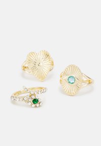 Três anéis de ouro: um com forma de coração e textura radial, outro com um motivo de olho apresentando uma pedra azul, e um terceiro com strass e uma pedra verde.