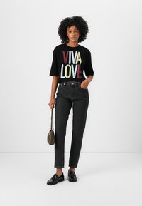 Juodas marškinėlis su spalvingu "VIVA LOVE" spaudiniu, derinamas su tamsiomis džinsomis ir juodais loafers. Modelis laiko mažą rudą rankinę.