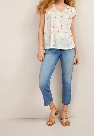 Vrouw draagt een witte top met korte mouwen met sterrenprint, lichtblauwe cropped jeans en beige sandalen met parelversieringen.