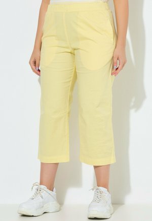 Frau trägt lockere gelbe Cropped-Hosen und weiße klobige Sneakers, steht vor weißem Hintergrund.