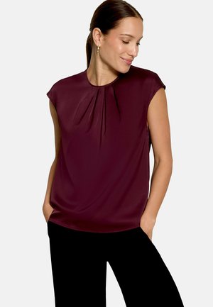Femme aux cheveux attachés portant des boucles d'oreilles créoles en or, un chemisier à manches courtes rouge foncé et un pantalon noir, les mains dans les poches.