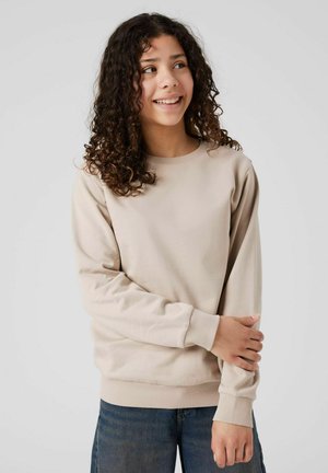 Jeune personne avec des cheveux bouclés portant un sweat-shirt beige et un jean bleu, souriant et regardant sur le côté les bras croisés.