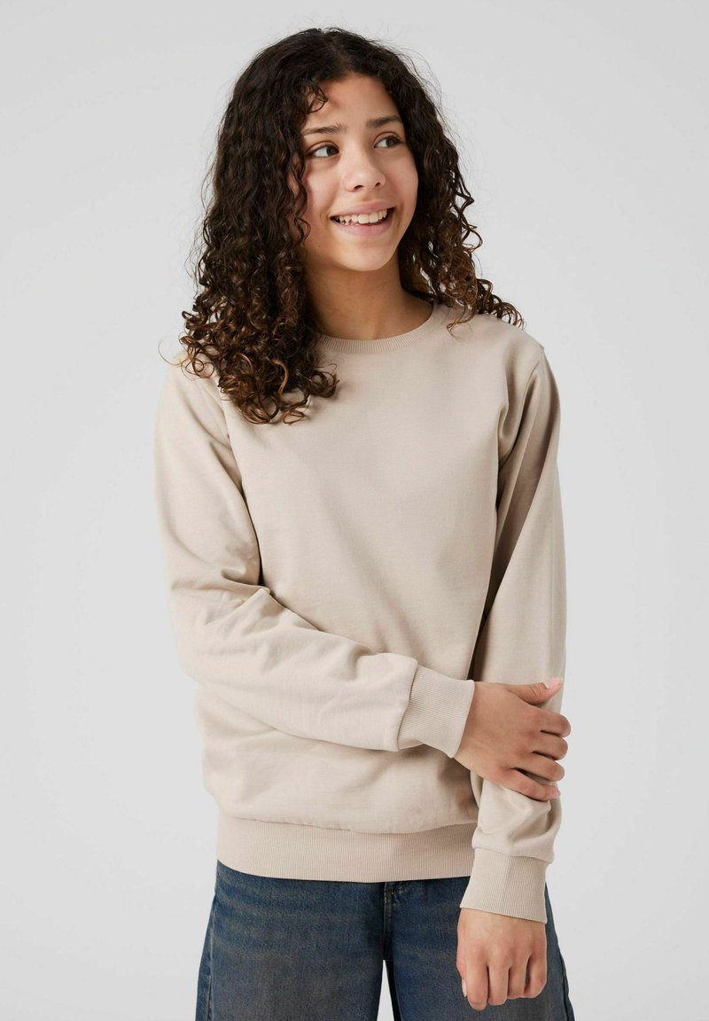 Giovane con capelli ricci che indossa una felpa beige e jeans blu, sorridendo e guardando di lato con le braccia incrociate.