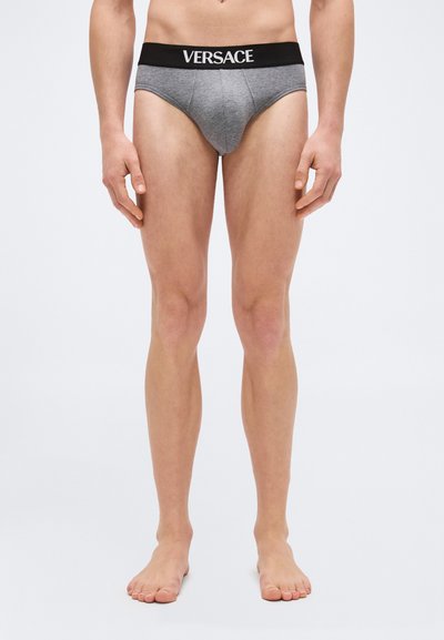 Torso et jambes d'un homme portant un slip gris Versace avec une ceinture noire, debout pieds nus sur un fond clair uni.