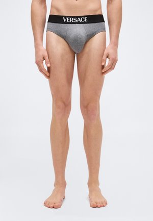 Άνδρας με κορμό και πόδια, φοράει γκρι εσώρουχα Versace με μαύρη ζώνη στη μέση, όρθιος ξυπόλητος πάνω σε απλό ανοιχτό φόντο.