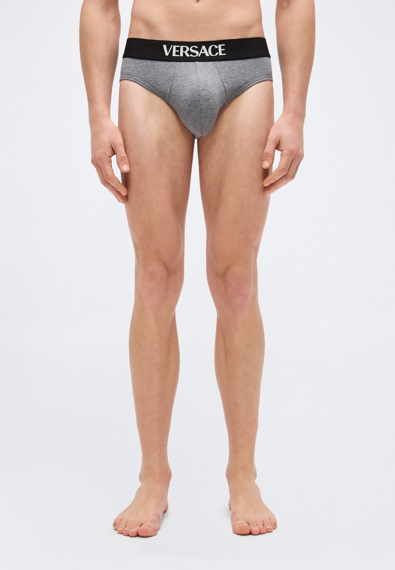 Άνδρας με κορμό και πόδια, φοράει γκρι εσώρουχα Versace με μαύρη ζώνη στη μέση, όρθιος ξυπόλητος πάνω σε απλό ανοιχτό φόντο.