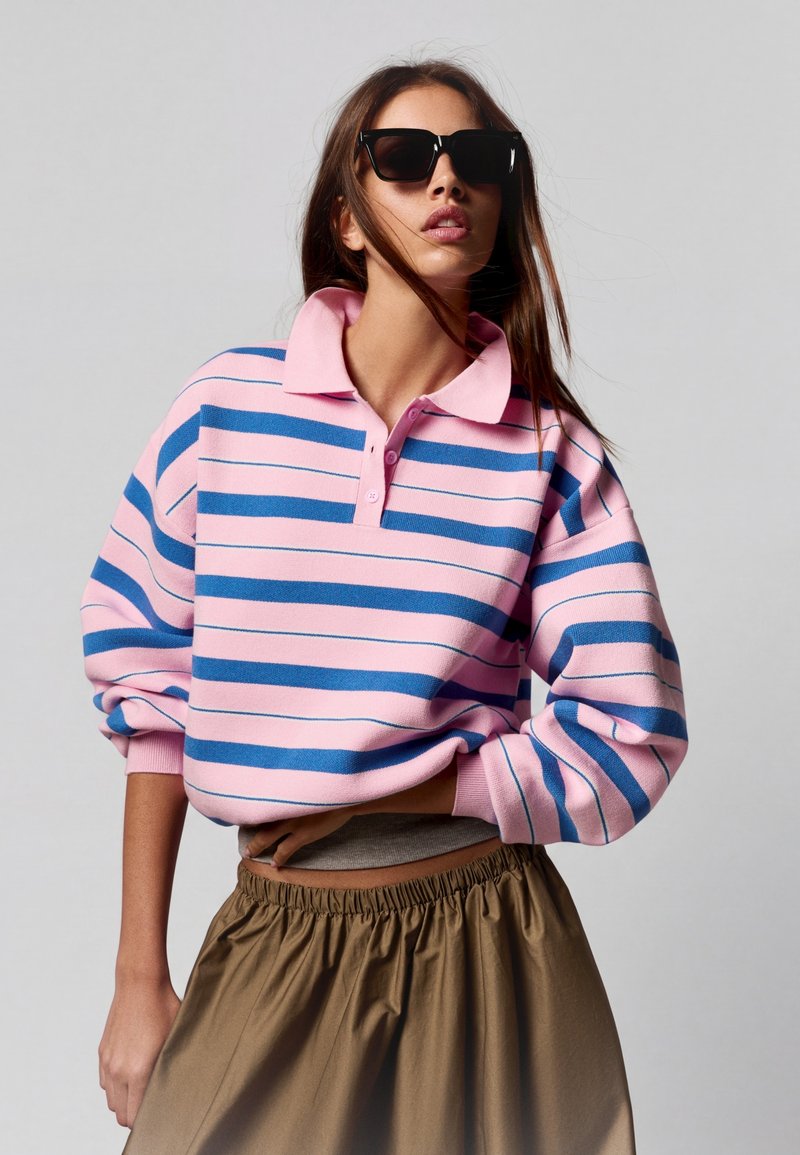 Femme portant des lunettes de soleil noires, un polo à manches longues rayé rose et bleu, et un pantalon marron taille haute sur un fond uni.