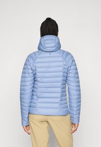 Osoba s pletenicama nosi svijetloplavu podstavljenu jaknu s kapuljačom i bež hlače, okrenuta leđima prema jednoličnoj pozadini.