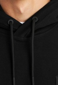 Primo piano del collo e della parte superiore del petto di una persona che indossa una felpa nera con cordini visibili e una camicia bianca sotto.